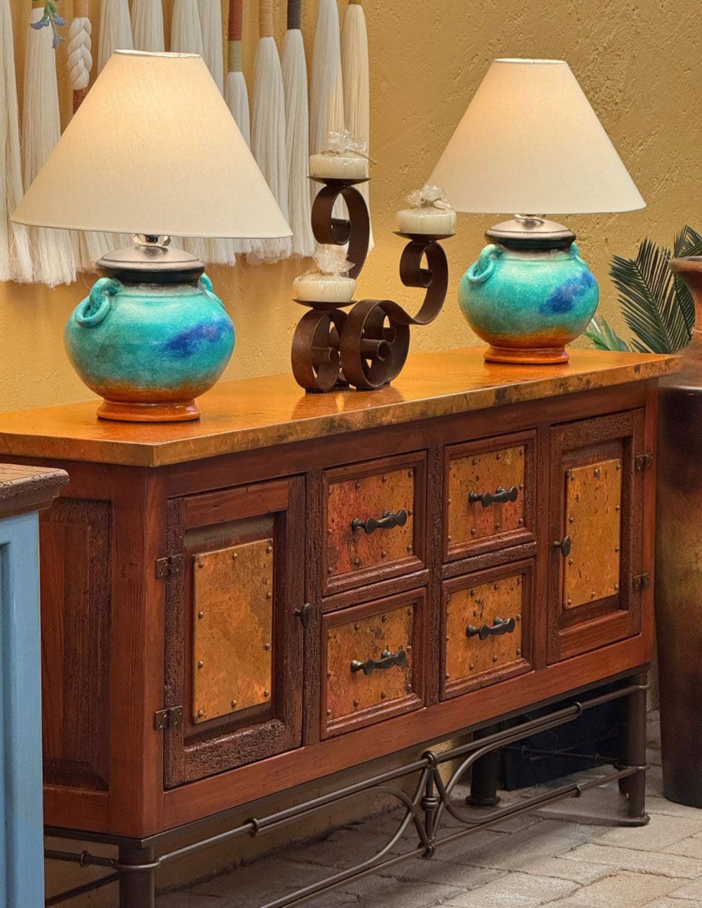 A wooden sideboard with copper-panelled doors on a wrought iron base, two turquoise glazed lamps and a carved candleholder on top