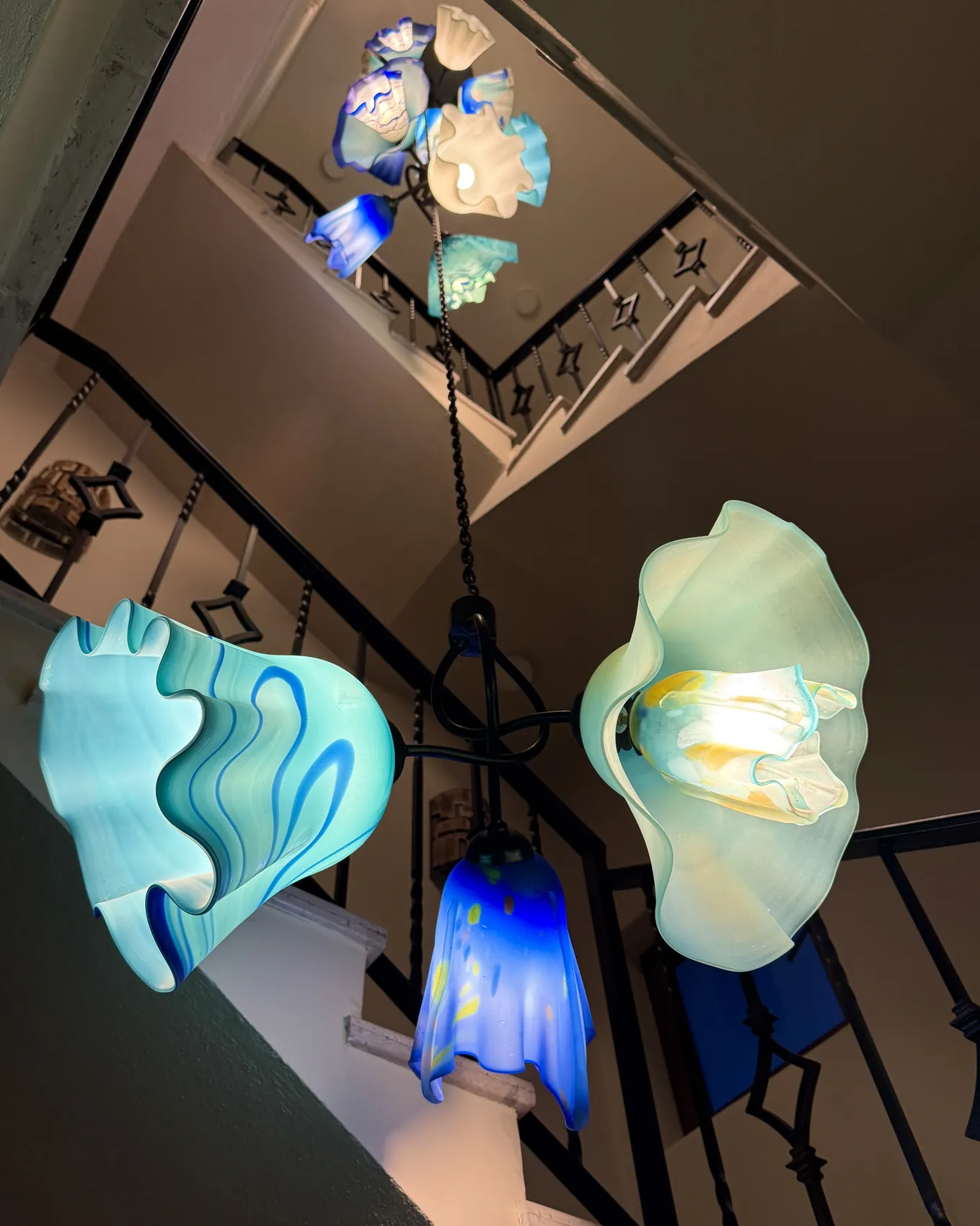 Blown glass shades in turquoise and blue hanging on ironwork, seen from below as they drop through an open stairwell
