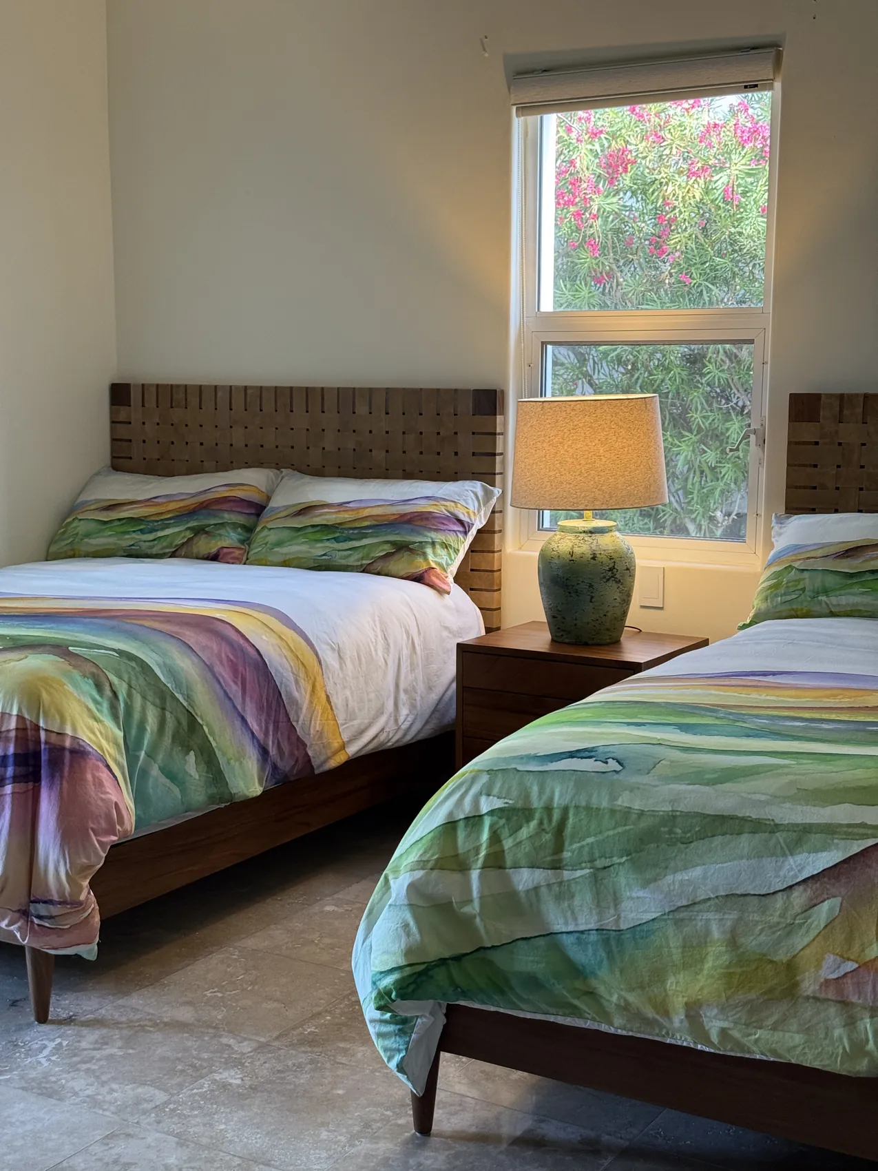 A parota bed with a woven leather headboard beside a window, made up in green and white striped linens, a glazed lamp on the nightstand