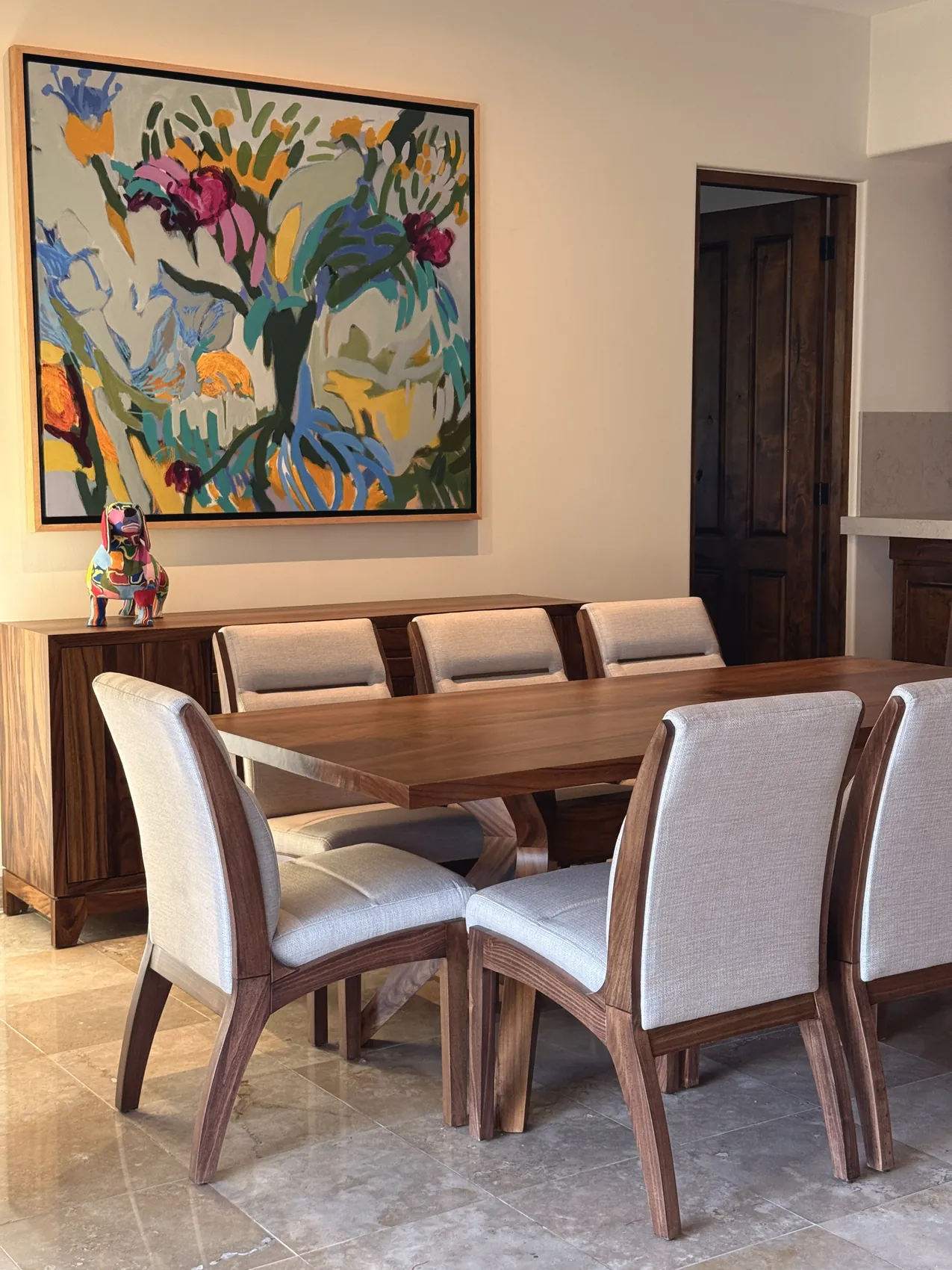 A long parota dining table with pale upholstered chairs on a travertine floor, a large floral canvas on the wall behind it