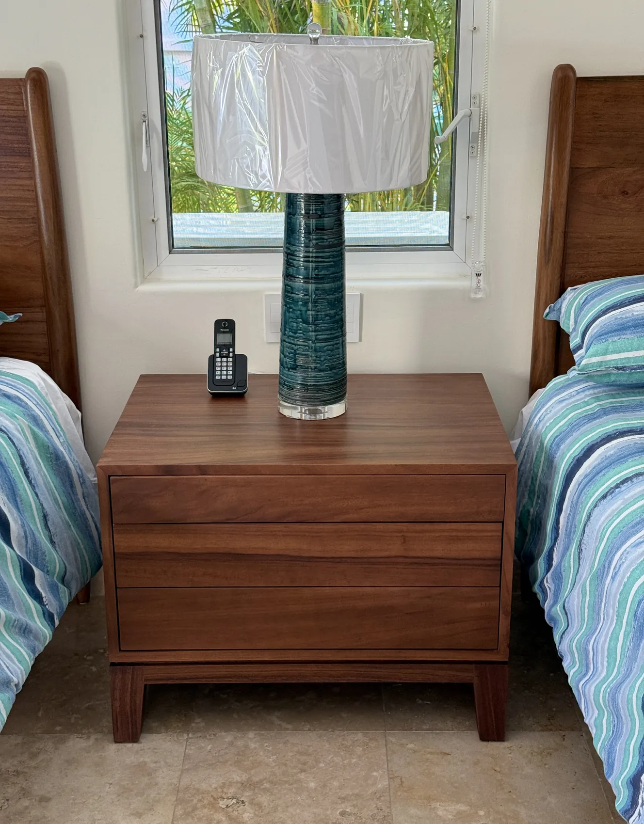 A parota nightstand with flush drawers standing between two beds under a window, a tall glazed lamp on top