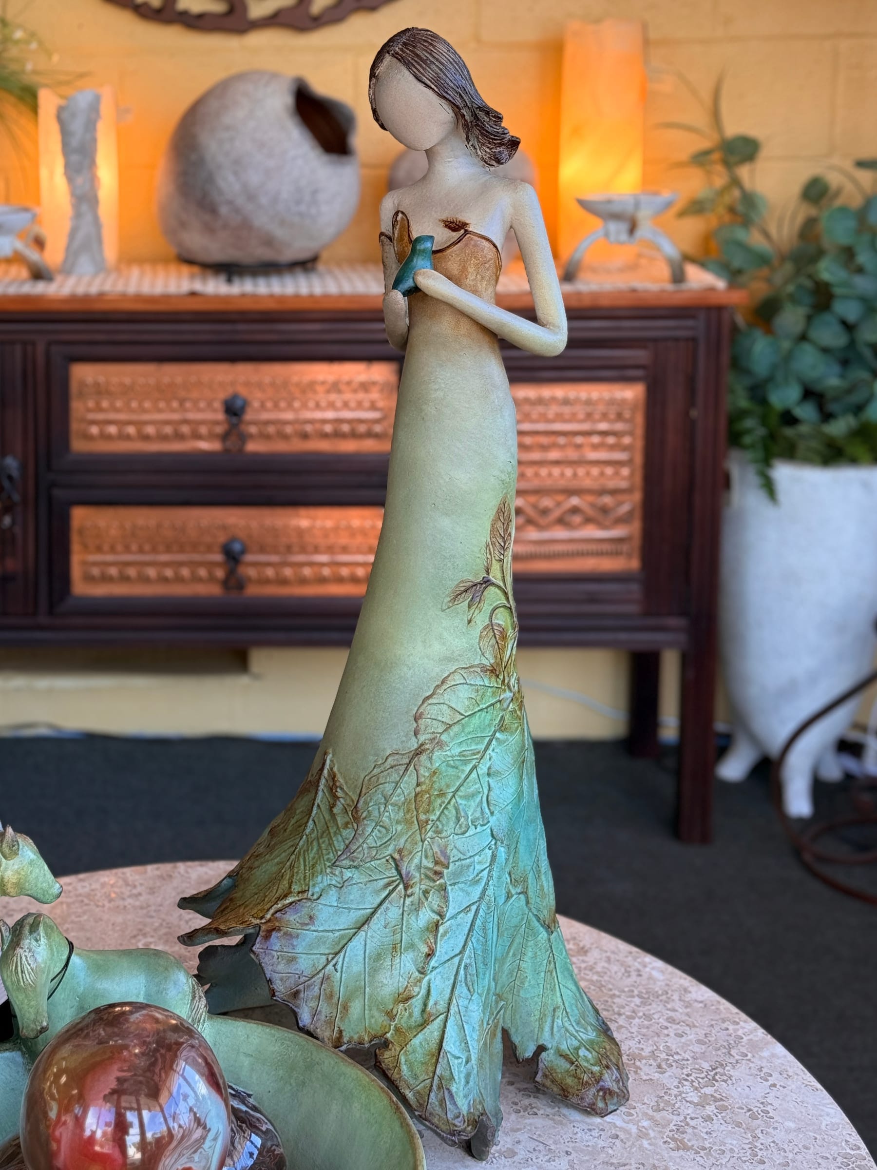 A tall sculpture of a woman in a leaf gown holding a bird, on a stone table in the Lake of the Ozarks showroom, with a carved console behind